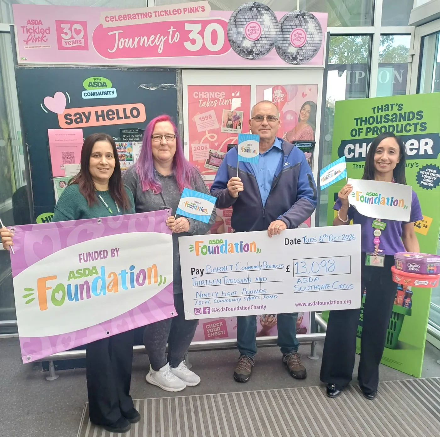 Four people holding Asda Foundation flags, a Funded by Asda Foundation banner and a £13,098 cheque in front of a Tickled Pink display at the entrance of Asda Southgate Circus