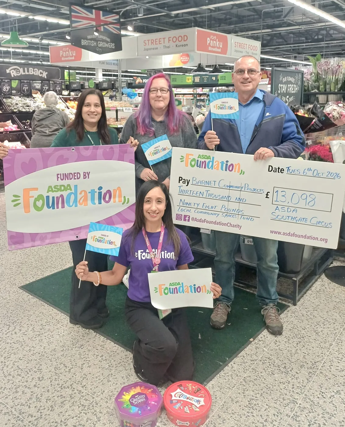 Four people with Asda Foundation flags, a banner and a £13,098 cheque in Asda Southgate Circus, with tins of Quality Street and Celebrations on the floor in front