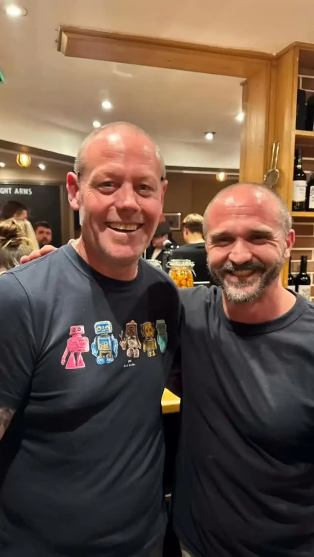 Two smiling men inside the bar of the Sebright Arms on its opening night, with drinkers and the bar behind them.